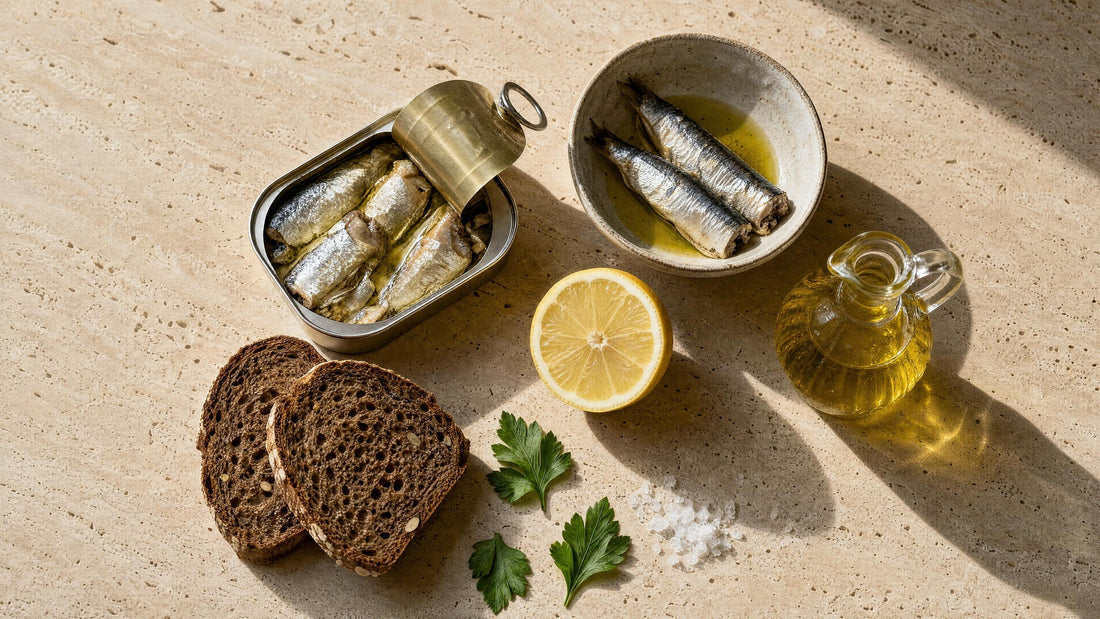 Geöffnete Sardinendose neben einer Schale mit Sardinen, Zitronenhälfte, Olivenöl in einer kleinen Karaffe und Vollkornbrot auf Travertin