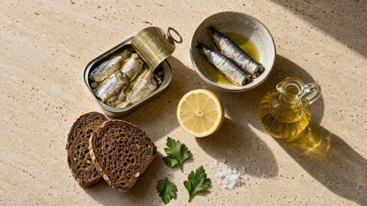 Geöffnete Sardinendose neben einer Schale mit Sardinen, Zitronenhälfte, Olivenöl in einer kleinen Karaffe und Vollkornbrot auf Travertin
