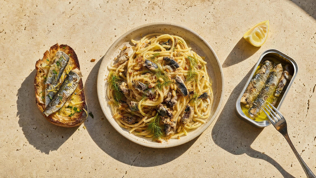Geröstetes Sauerteigbrot mit Sardinen, Zitrone und Petersilie, daneben Pasta mit Sardinen und Fenchel und eine geöffnete Sardinendose auf Travertin